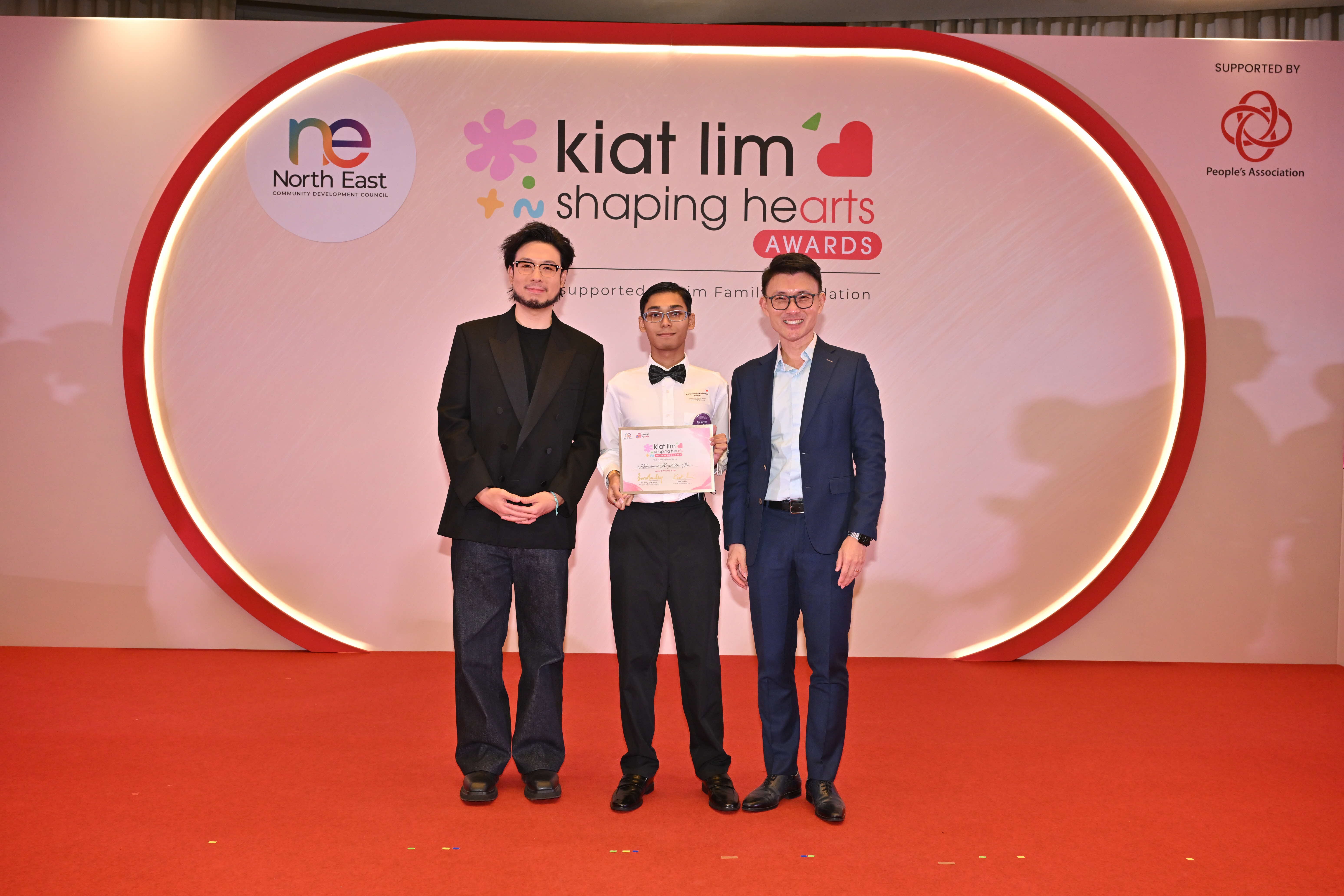 Award recipient standing on stage with the presenter, holding their certificate and posing for a group photo during the award ceremony, with a backdrop reading ‘Kiat Lim-Shaping Hearts Awards'.